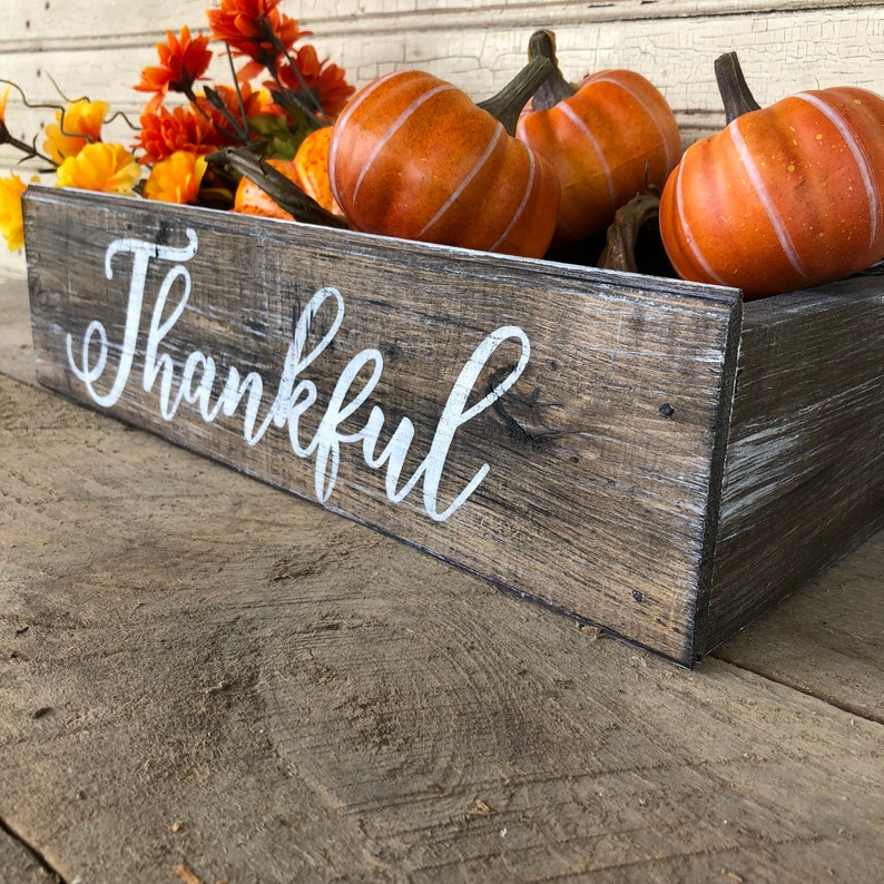 May include: A rustic wooden box with the word "Thankful" painted in white on the front. The box is filled with orange and yellow fall-themed decorations, including pumpkins and flowers.
