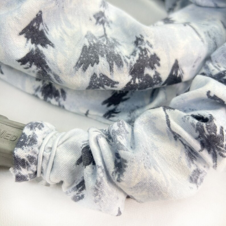 Pu&ograve; includere: Un elastico per capelli in tessuto azzurro con una stampa grigio scuro di alberi sempreverdi. L'elastico &egrave; arricciato con una fascia elastica e copre parzialmente un accessorio per capelli in plastica grigia con il testo visibile MED.
