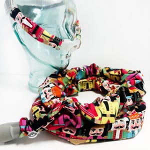 May include: A colorful fabric tube with a Nutcracker pattern, designed to cover a CPAP hose. The tube is made of soft, stretchy fabric and features a variety of colors, including red, green, blue, yellow, and black. The fabric is printed with a repeating pattern of Nutcracker figures.