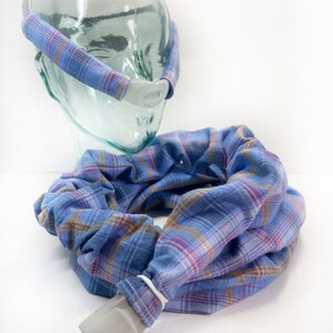 May include: A blue and orange plaid fabric headband with a white plastic piece on the front. The headband is wrapped around a glass head form. A matching blue and orange plaid fabric tube is lying on the table.