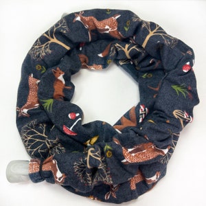 May include: A dark gray infinity scarf with a repeating pattern of brown deer, foxes, trees, and mushrooms. The scarf is made of soft fabric and has a gray clasp. The design is whimsical and nature-themed.