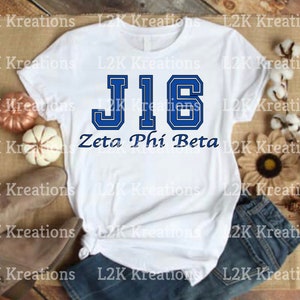 May include: White t-shirt with a blue and white varsity style "J16" graphic and the text "Zeta Phi Beta" printed on the front.