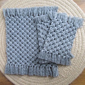 May include: A set of two gray macrame placemats with matching coasters. The placemats and coasters are made of woven cotton and have a diamond pattern. Each piece has a fringe trim along the edges. The placemats and coasters are on a beige woven surface.