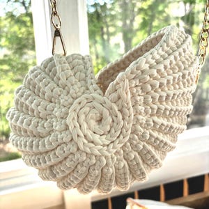 May include: Cream-colored, handmade crochet basket in a nautilus shell design, suspended by a gold chain. The basket features a spiral center and a textured, ridged exterior. The background shows a window with a view of green foliage.