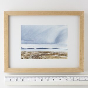 May include: A framed watercolor painting of a seascape with a cloudy sky and a distant shoreline. The painting is in a light wood frame.