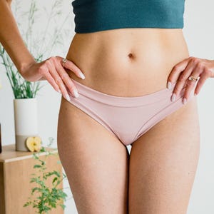 May include: A teal crop top and pale pink underwear. The top has a ruched front and a scoop neckline. The underwear is a low-rise style. The person is wearing rings on their fingers.