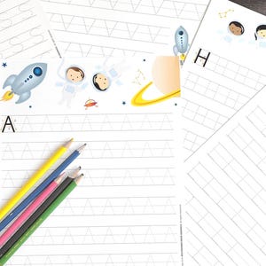 以下が含まれることがあります： 宇宙をテーマにしたイラストが描かれた、白い罫線入りの紙。宇宙飛行士、惑星、ロケットなどが描かれています。手書き練習用の文字が書かれており、「S」、「A」、「V」、「H」、「L」の文字が見えます。色鉛筆も写っています。
