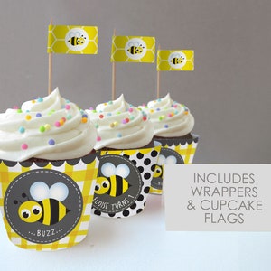 May include: Three yellow and black bee-themed cupcake wrappers with a black and white polka dot design. The wrappers have a cute bee illustration and the text "...Buzz..." and "Eloise Turns 1".  The cupcakes are topped with white frosting and colorful sprinkles.  Three yellow and black bee-themed cupcake flags with a honeycomb design are included.  The text "INCLUDES WRAPPERS & CUPCAKE FLAGS" is printed on a white card.