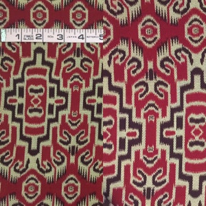 May include: Red and beige fabric with a geometric pattern. The fabric has a woven texture and features a repeating design of stylized figures. The fabric is measured with a tape measure that reads "Fiber Glass" and "Japan Butterfly".