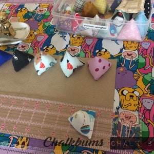 May include: A close-up of eight triangular fabric weights, each with a different pattern. The weights are arranged in a row on a brown surface with a ruler and the words "Chalkbuns" and "Chalk Bags" visible.