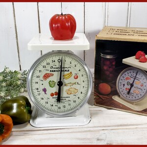 May include: White vintage kitchen scale with a black needle and a white dial that reads "The American Family Scale" and has a picture of various fruits, vegetables, and meat. The scale is on a white wooden surface with a red apple on top of the scale.