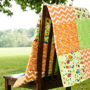 以下が含まれることがあります： オレンジ、グリーン、ホワイトの生地で作られたパッチワークキルト。キルトは木製の椅子にかけられています。