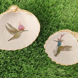 Könnte beinhalten: Zwei dekorative Muschelschalen mit Kolibri-Illustrationen. Die Muscheln sind cremefarben mit goldfarbenen Rändern. Ein Kolibri hat eine rosa Blume, der andere blaue und grüne Federn.