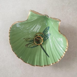 May include: A pale green seashell-shaped trinket dish with a gold trim. The dish has a dragonfly design and holds two gold rings. This decorative dish is suitable for storing jewellery or small items.