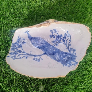 May include: A decorative seashell dish featuring a blue peacock design. The peacock is depicted on a branch with blue leaves. The shell's rim is trimmed with gold. The dish is on a green surface.