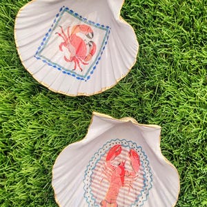 May include: Two white seashell-shaped dishes with gold trim, each featuring a different sea creature illustration. One has a red crab design, the other a red lobster. The dishes are on a bed of green grass.