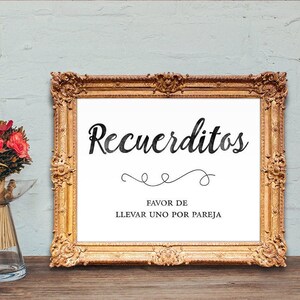 May include: A gold framed sign with the text "Recuerditos" in black cursive lettering. Below the text is "Favor de Llevar Uno Por Pareja" in black lettering.