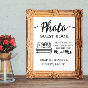 以下が含まれることがあります： インスタントカメラの白黒イラストと黒い文字が書かれた白い看板。看板には「Photo Guest Book Leave a photo and your wishes for the new Mr. and Mrs. Snap it, shake it, sign it, peg it.」と書いてあります。
