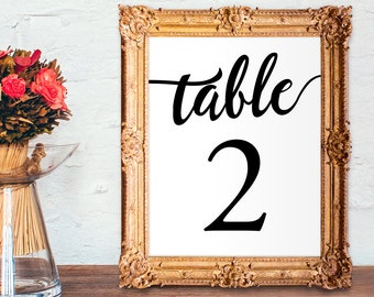 May include: A gold-framed table number sign with the word "table" and the number "2" in black calligraphy. The sign is on a wooden surface.