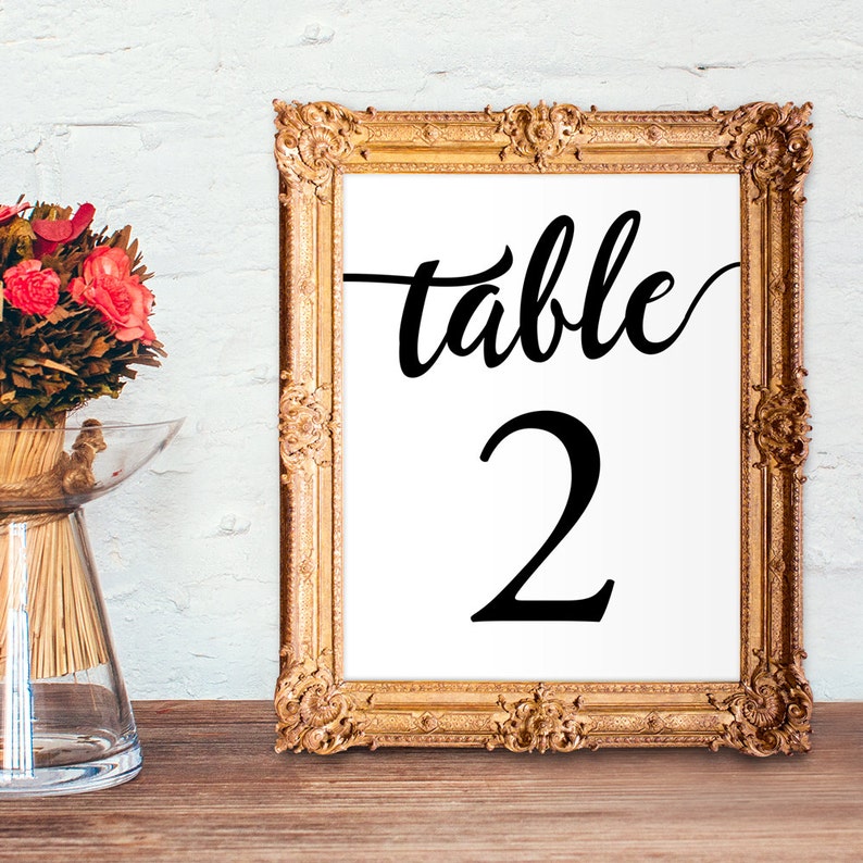 May include: A gold-framed table number sign with the word "table" and the number "2" in black calligraphy. The sign is on a wooden surface.