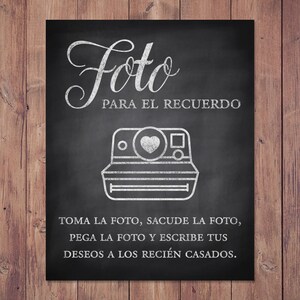 May include: Black chalkboard sign with white text and a drawing of a vintage instant camera. The text reads "Foto PARA EL RECUERDO" and instructions in Spanish. The sign is set against a wooden background.
