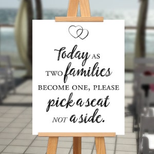 May include: A white sign with black text and two intertwined hearts at the top. The text reads "Today as two families become one, please pick a seat not a side."