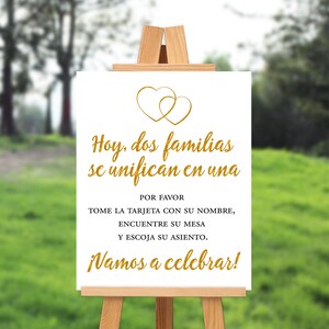 May include: A white sign with gold text and two gold hearts at the top. The sign reads "Hoy, dos familias se unifican en una. POR FAVOR TOME LA TARJETA CON SU NOMBRE, ENCUENTRE SU MESA Y ESCOJA SU ASIENTO. ¡Vamos a celebrar!"