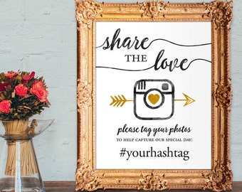 Puede incluir: Un cartel blanco en un marco dorado con el texto "share the love" en escritura negra. Una flecha dorada apunta a un icono de cámara de Instagram negro y dorado con un corazón dorado. El texto "please tag your photos to help capture our special day: #yourhashtag" está debajo del icono.