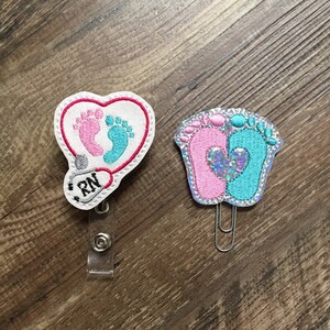 May include: Two embroidered badge clips, one with a heart shape and the words "RN" and the other with a pair of baby footprints, one pink and one blue.