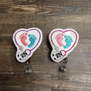 May include: Two white heart-shaped embroidered badge holders with a stethoscope and baby footprints. The badge holders have a clear plastic retractable reel and say "RN" on them.