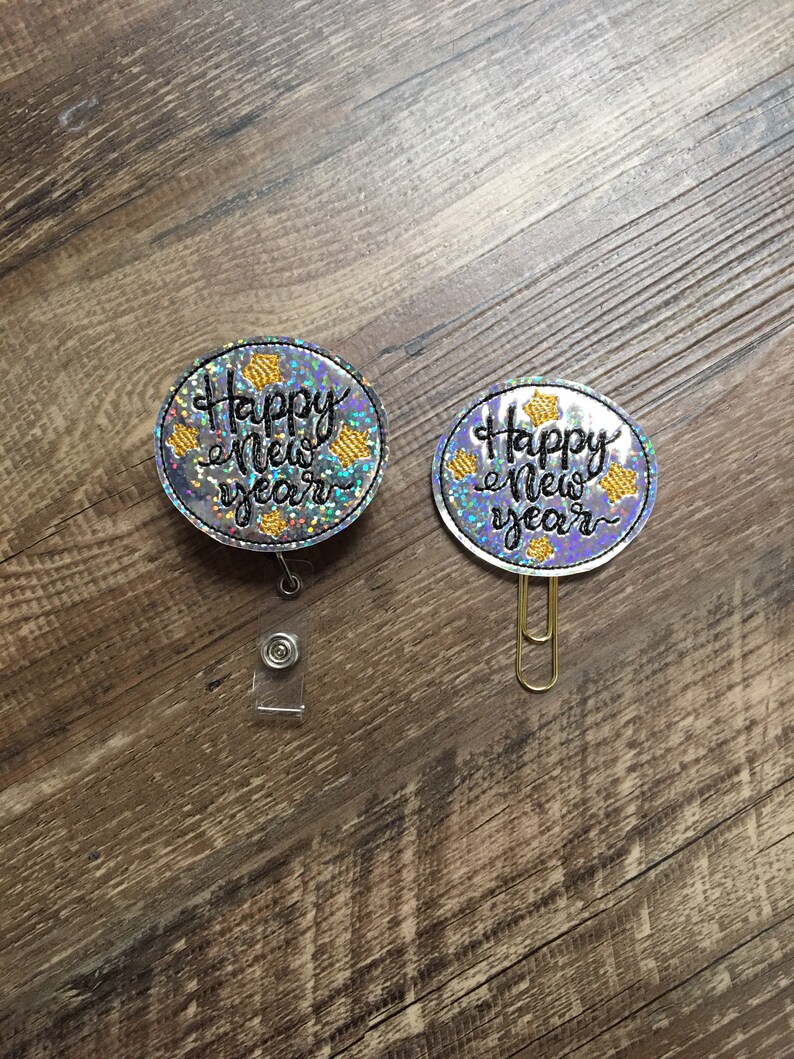 May include: Two silver glitter circle badges with gold stars and black text that reads "Happy New Year". One badge has a clear plastic badge holder attached and the other has a gold paper clip attached.