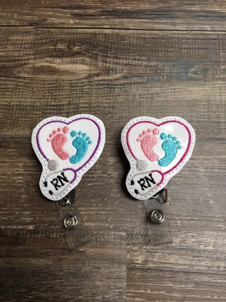 May include: Two white heart-shaped embroidered badge holders with a stethoscope and baby footprints. The badge holders have a clear plastic retractable reel and say "RN" on them.