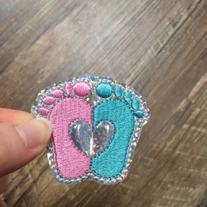 May include: A pink and blue baby footprint applique with a silver glitter heart in the center. The applique is on a clear background.