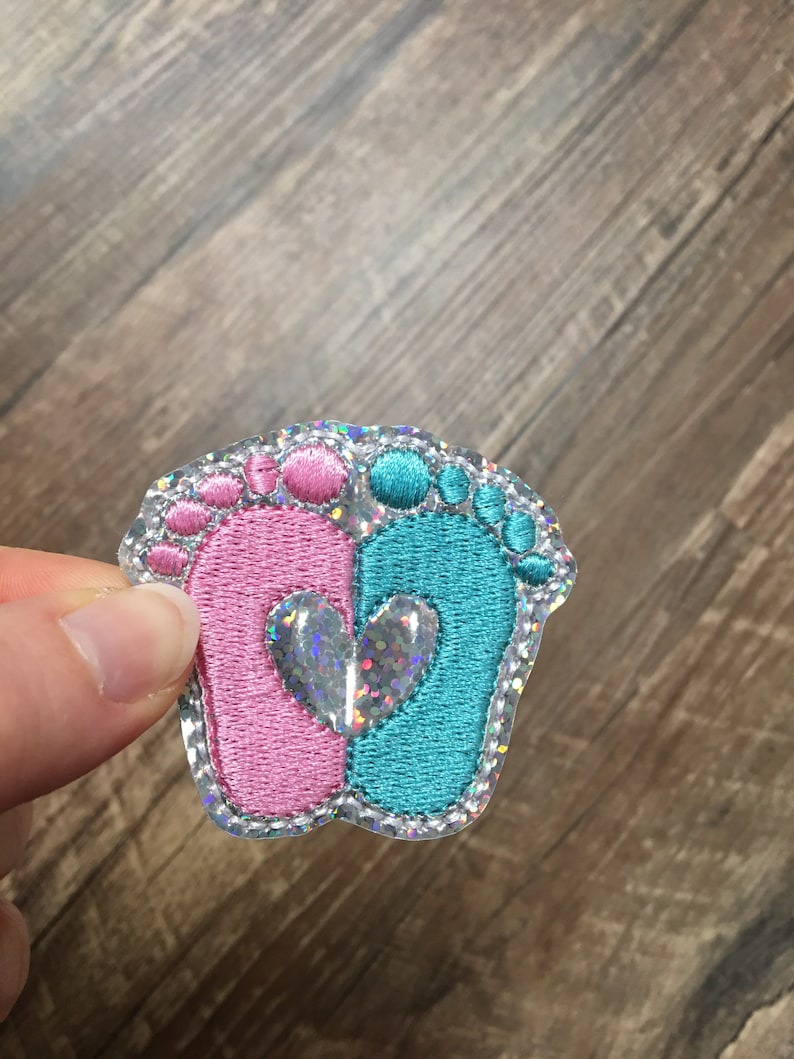 May include: A pink and blue baby footprint applique with a silver glitter heart in the center. The applique is on a clear background.