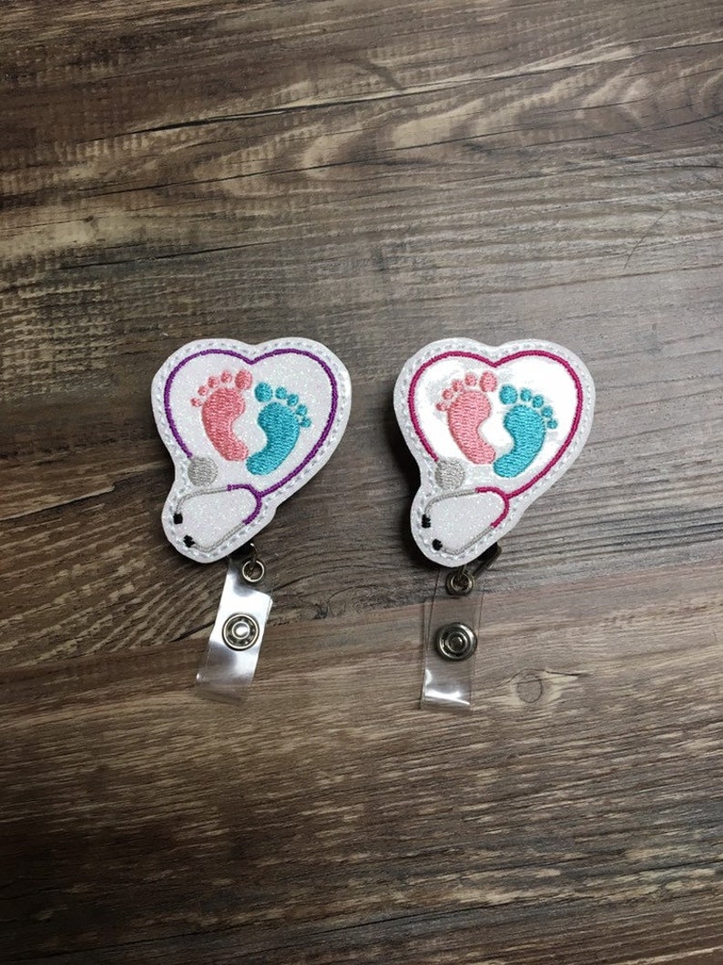 May include: Two white heart-shaped badge holders with a stethoscope design and pink and blue baby footprints. The badge holders have clear plastic straps.