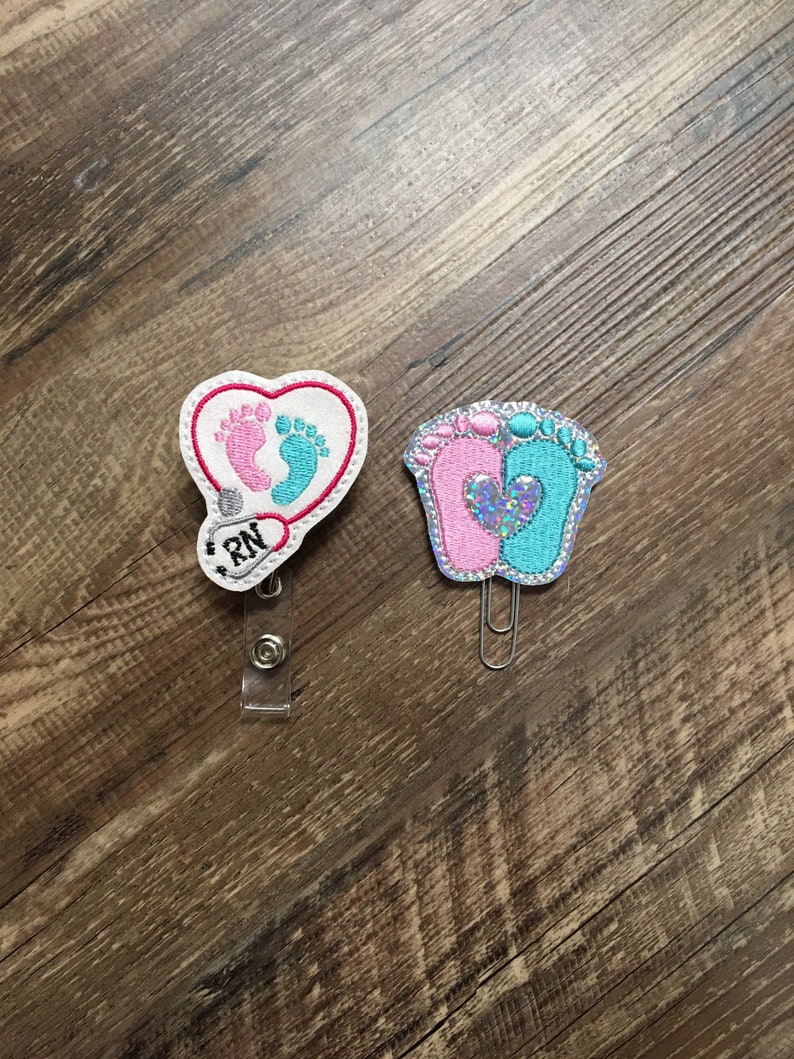 May include: Two embroidered badge clips, one with a heart shape and the words "RN" and the other with a pair of baby footprints, one pink and one blue.