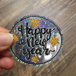May include: A round, silver, iridescent patch with black embroidery that reads "Happy New Year" and gold star accents.