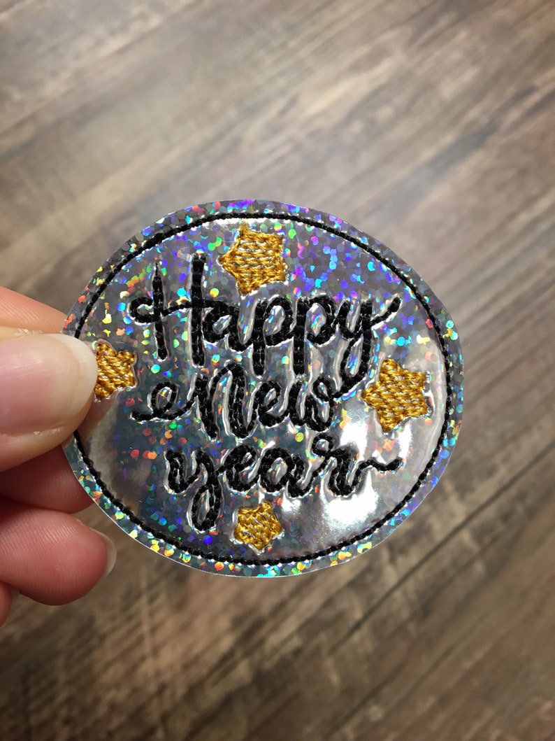 May include: A round, silver, iridescent patch with black embroidery that reads "Happy New Year" and gold star accents.