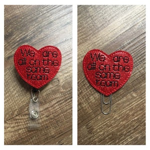 May include: Two red glitter heart-shaped badges with the text "We are all on the same team" in black. One badge is attached to a clear plastic badge holder with a clip, the other is attached to a silver paper clip.