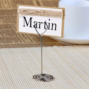Może przedstawiać: Srebrny stojak na wizyt&oacute;wki w kształcie serca z podstawą spiralną. Brązowa wizyt&oacute;wka z wstążką jutową i imieniem "Martin" jest wyświetlana w stojaku.
