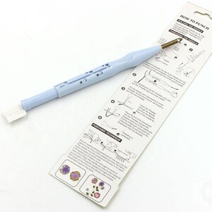May include: A light blue plastic tool with a white handle and a gold needle. The tool is used for punching holes in fabric for crafts. The packaging includes instructions on how to use the tool.