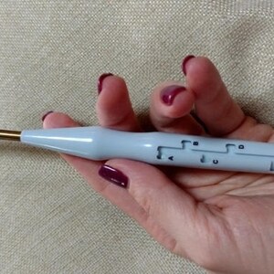 May include: A white and gold crochet hook with a built-in yarn needle. The hook has a clear plastic handle with markings for different sizes.