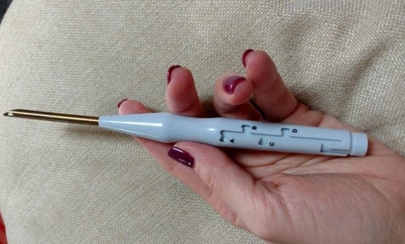 May include: A white and gold crochet hook with a built-in yarn needle. The hook has a clear plastic handle with markings for different sizes.
