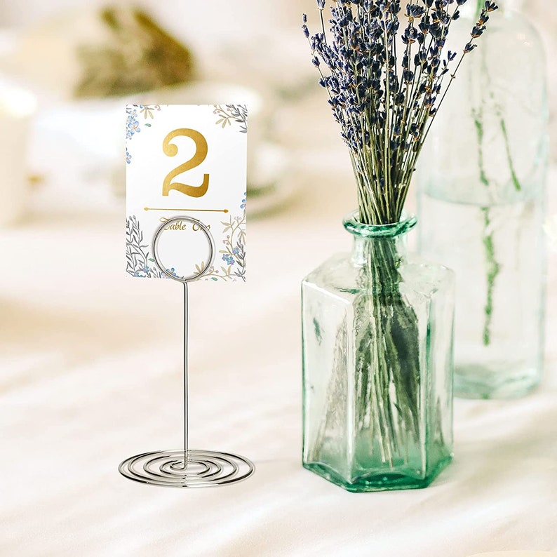 Pode incluir: Um porta-n&uacute;meros de mesa prateado com um cart&atilde;o branco com um design floral e o n&uacute;mero "2" em letras douradas. O cart&atilde;o tamb&eacute;m diz "Table One".