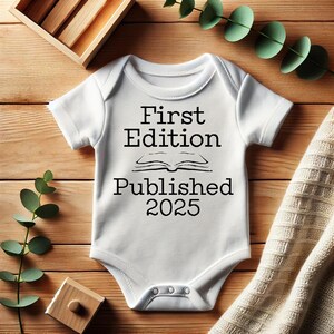 May include: White baby onesie with the text "First Edition Published 2025" in black. The onesie has short sleeves and a snap closure. The image is on a wooden surface with decorative elements.