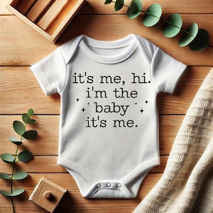 May include: White baby onesie with black text that reads "it's me, hi. i'm the baby it's me."