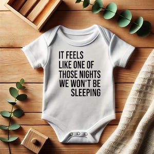 以下が含まれることがあります： 「IT FEELS LIKE ONE OF THOSE NIGHTS WE WON'T BE SLEEPING」の黒い文字が入った白いベビーボディスーツ。木製の表面に置かれ、装飾的な要素があります。