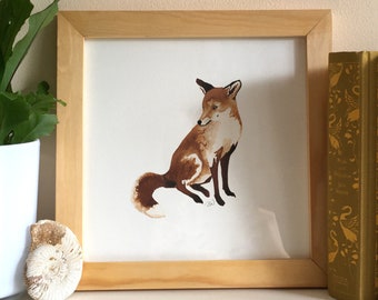 Puede incluir: Una impresión enmarcada de una pintura de acuarela de un zorro sentado sobre un fondo blanco. El marco es de madera clara y tiene un diseño simple. La impresión se muestra sobre una superficie blanca con una planta en maceta y algunos libros.