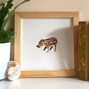 May include: A framed watercolour painting of a brown and white wild boar on a white background. The frame is made of light wood and is sitting on a white surface. There are three books behind the frame, two are brown and one is gold.