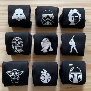 May include: Set of 10 black socks with white Star Wars character graphics. The characters include Darth Vader, a Stormtrooper, Chewbacca, R2-D2, a Rebel pilot, Yoda, Boba Fett, and a Mandalorian.